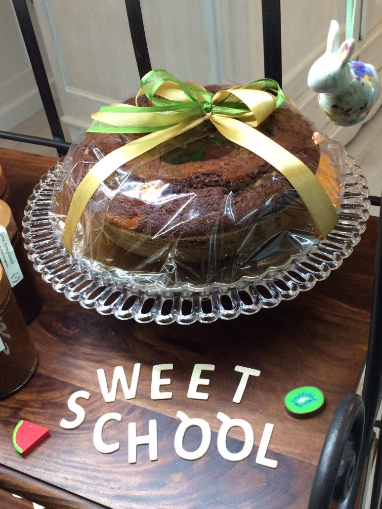 Ciambellone incartato con nastro giallo e verde su un'alzata di vetro, accanto ai vasetti di confettura