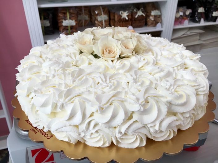 Torta ricoperta di ciuffi di panna montata con un mazzetto di rose bianche al centro, sul vassoio Aldemorisco