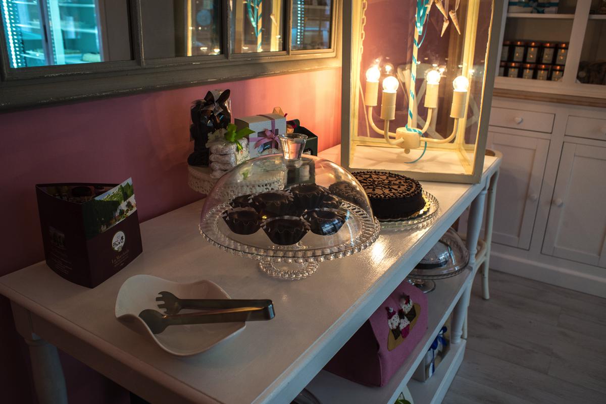 Interno del negozio Aldemorisco: alzata di vetro con tortine di cioccolato e crostata su un tavolo bianco, parete rosa e lanterna accesa